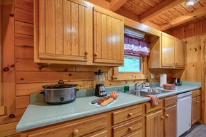 A wooden cabin kitchen with a stoneware crockpot, a blender, a sink, and a window with a plaid curtain.