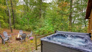 Ein Whirlpool auf einer Holzterrasse, umgeben von Stühlen und einer Feuerstelle, eingebettet in eine bewaldete Landschaft.
