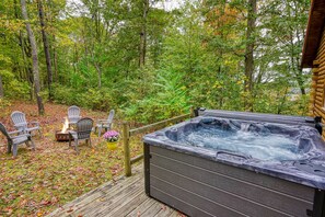 Ein Whirlpool auf einer Holzterrasse, umgeben von Stühlen und einer Feuerstelle, eingebettet in eine bewaldete Landschaft.