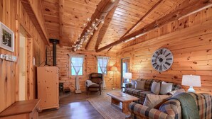 A cozy cabin interior with wooden walls, a large clock, a sofa, a chair, and a fireplace. - Cozy Casa Carezza Log Cabin, Logan, Ohio, Pet Friendly, Sleeps 6 (Logan)