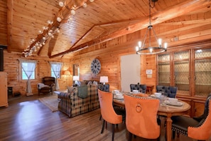 A cozy log cabin interior with a dining area, a living room with a sofa and chairs, and a large wall clock.