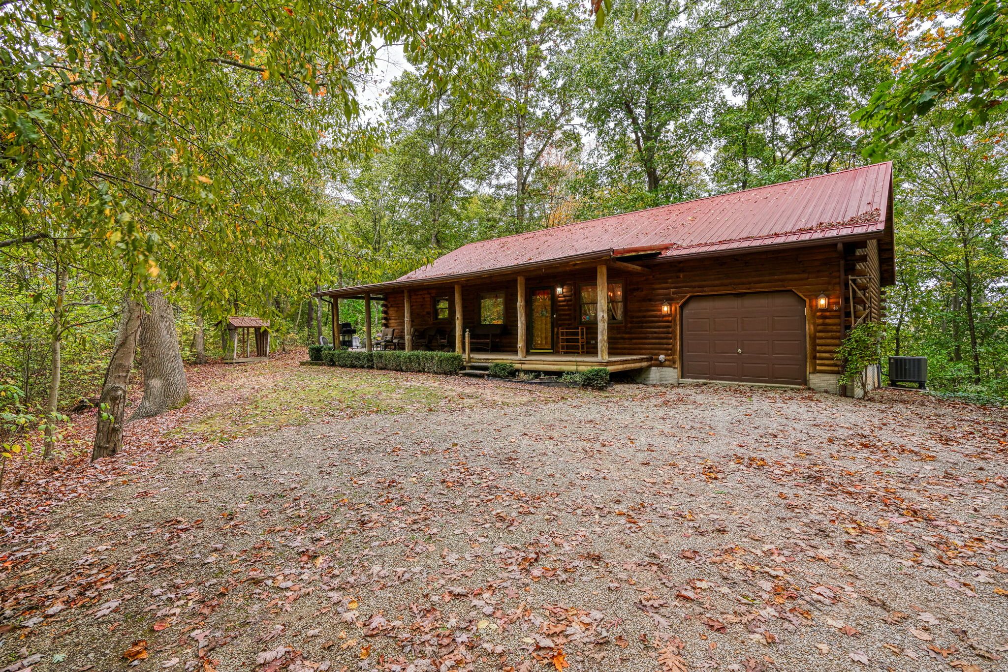 A rustic cabin with a covered porch, a garage, and a gravel driveway surrounded by trees.