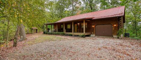 A rustic cabin with a covered porch, a garage, and a gravel driveway surrounded by trees.