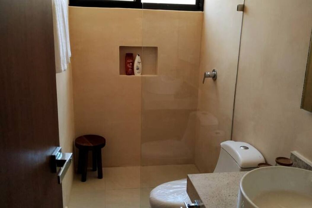 A small bathroom with a toilet, a sink, and a shower stall. There is a small stool and a soap dispenser on the wall.
