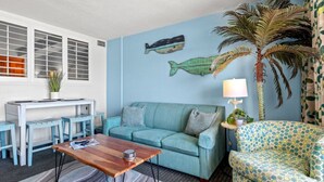 Ein Wohnzimmer mit einem blauen Sofa, einem Holztisch, einem gemusterten Sessel, einer Wanddekoration mit Fischen, einer hohen Zimmerpalme und einem kleinen Tisch mit einer Lampe.
