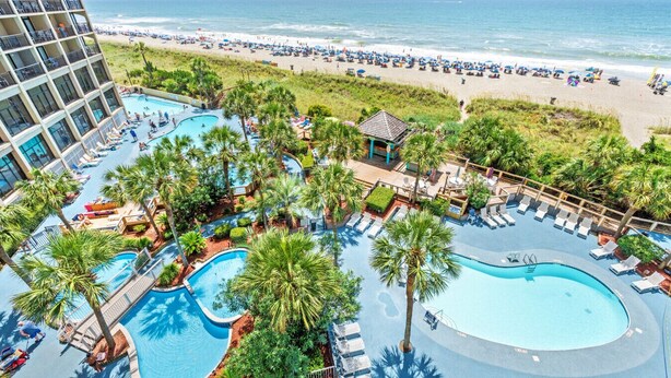 Ein Resort mit mehreren Schwimmbecken, umgeben von Palmen, einem Strand und einem Hochhaus.