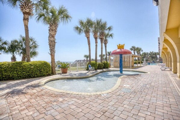 A paved area with a pool, palm trees, a fountain, and a building with arches.