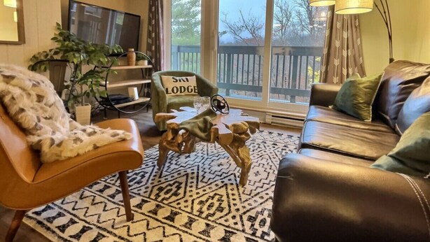 A living room with a leather sofa, a green armchair, a unique wooden coffee table, a television, and a large window with curtains.