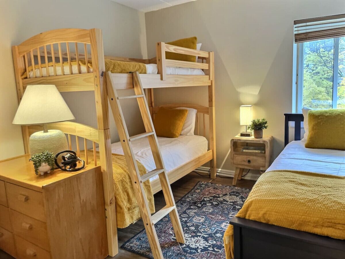 A bunk bed room with a desk, a small bedside table, a lamp, and a window with a view of trees.