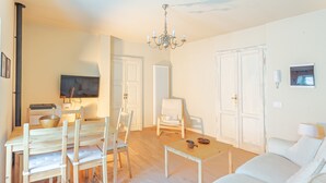 A living room with a white sofa, wooden dining table, and chairs. There is a television on the wall and a chandelier. - PM9        (Castello Cabiaglio)