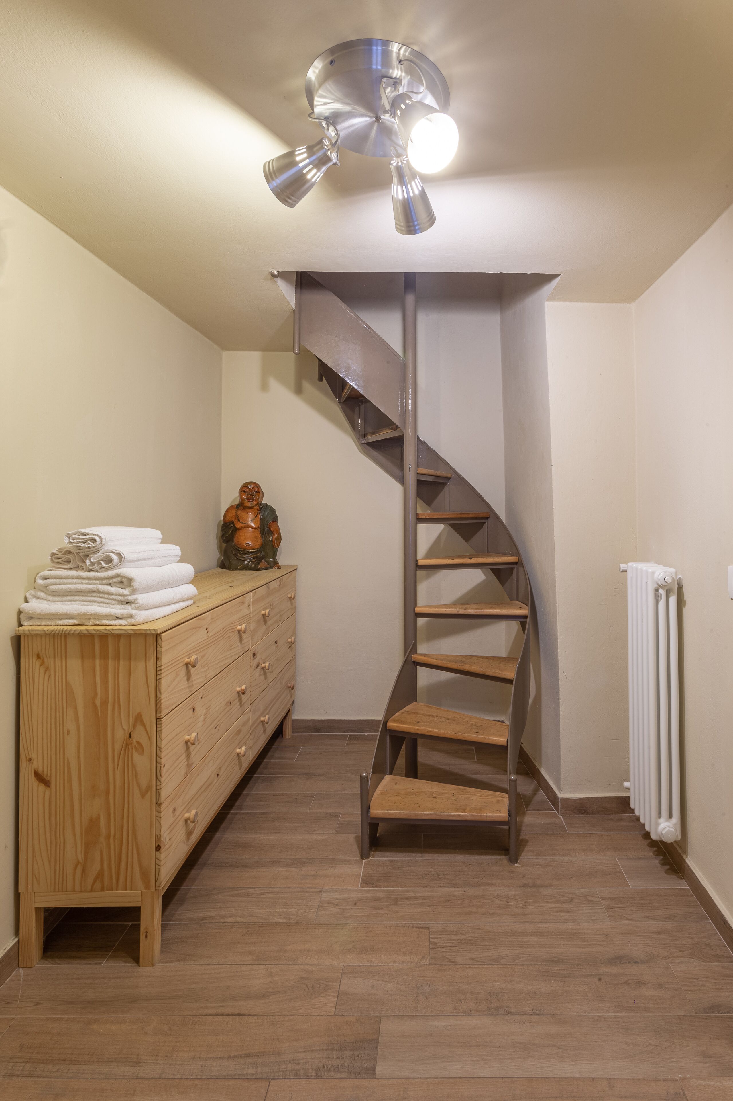 Un escalier en colimaçon dans une pièce dont le sol est recouvert de planches de bois, une commode en bois avec des serviettes pliées, et un radiateur fixé au mur.