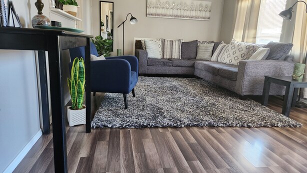 A living room with a dark wood floor, a blue armchair, a grey sectional sofa, a patterned rug, and a large mirror on the wall.