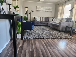 Una sala de estar con piso de madera oscura, un sillón azul, un sofá seccional gris, un tapete estampado y un espejo grande en la pared.