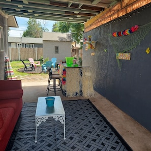 Une terrasse couverte avec une table et des chaises, un petit coin bar agrémenté d’une pancarte « Bienvenue », et une guirlande de fleurs colorée accrochée au mur.