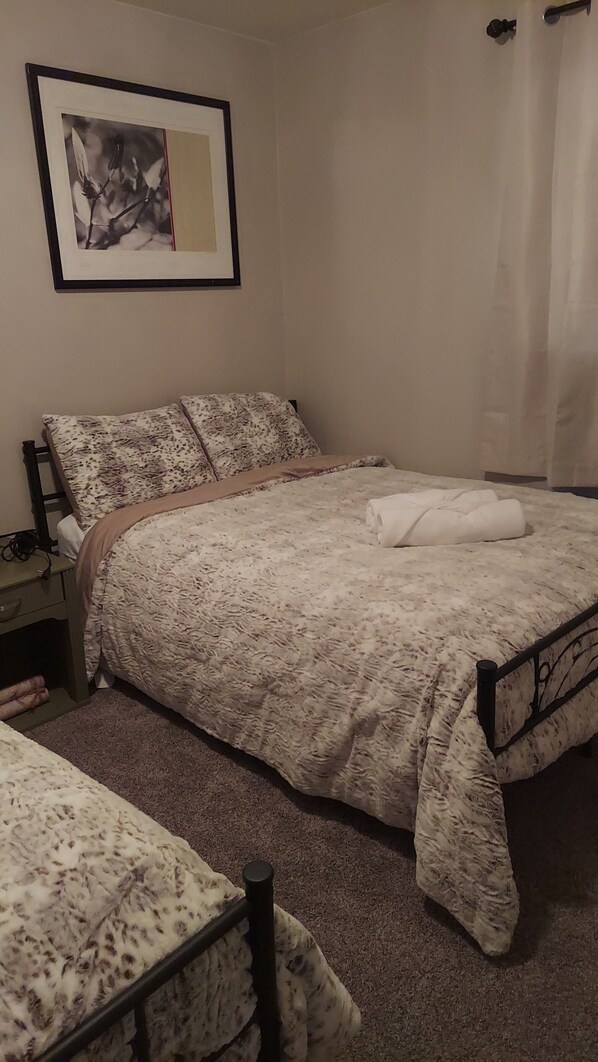 Un dormitorio con cama, mesita de noche, silla, un cuadro enmarcado en la pared y una ventana con cortinas.