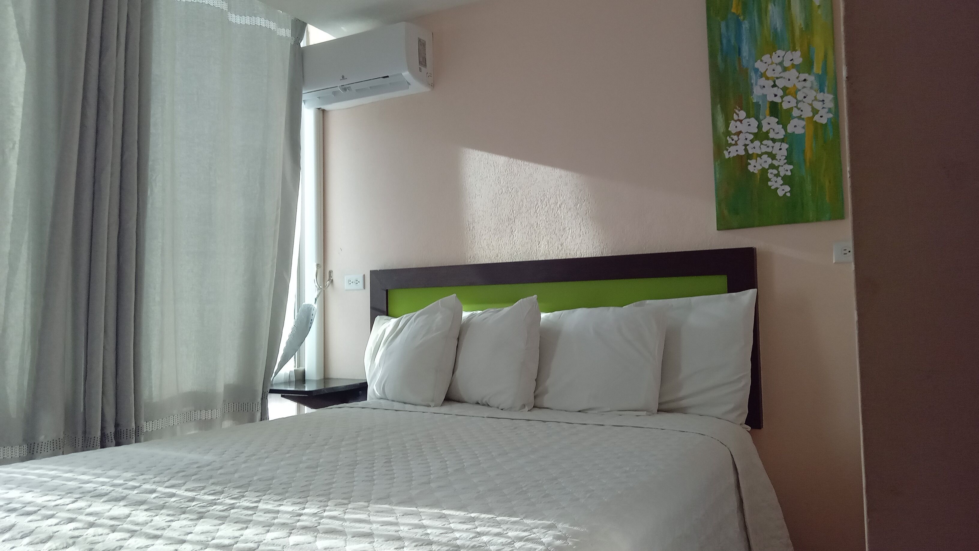 Una cama bien hecha con ropa blanca y un cabecero verde, una mesita de noche con una lámpara y un cuadro de flores en la pared.