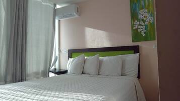 Een net opgemaakt bed met witte lakens en een groen hoofdeinde, een nachtkastje met een lamp, en een schilderij van bloemen aan de muur.