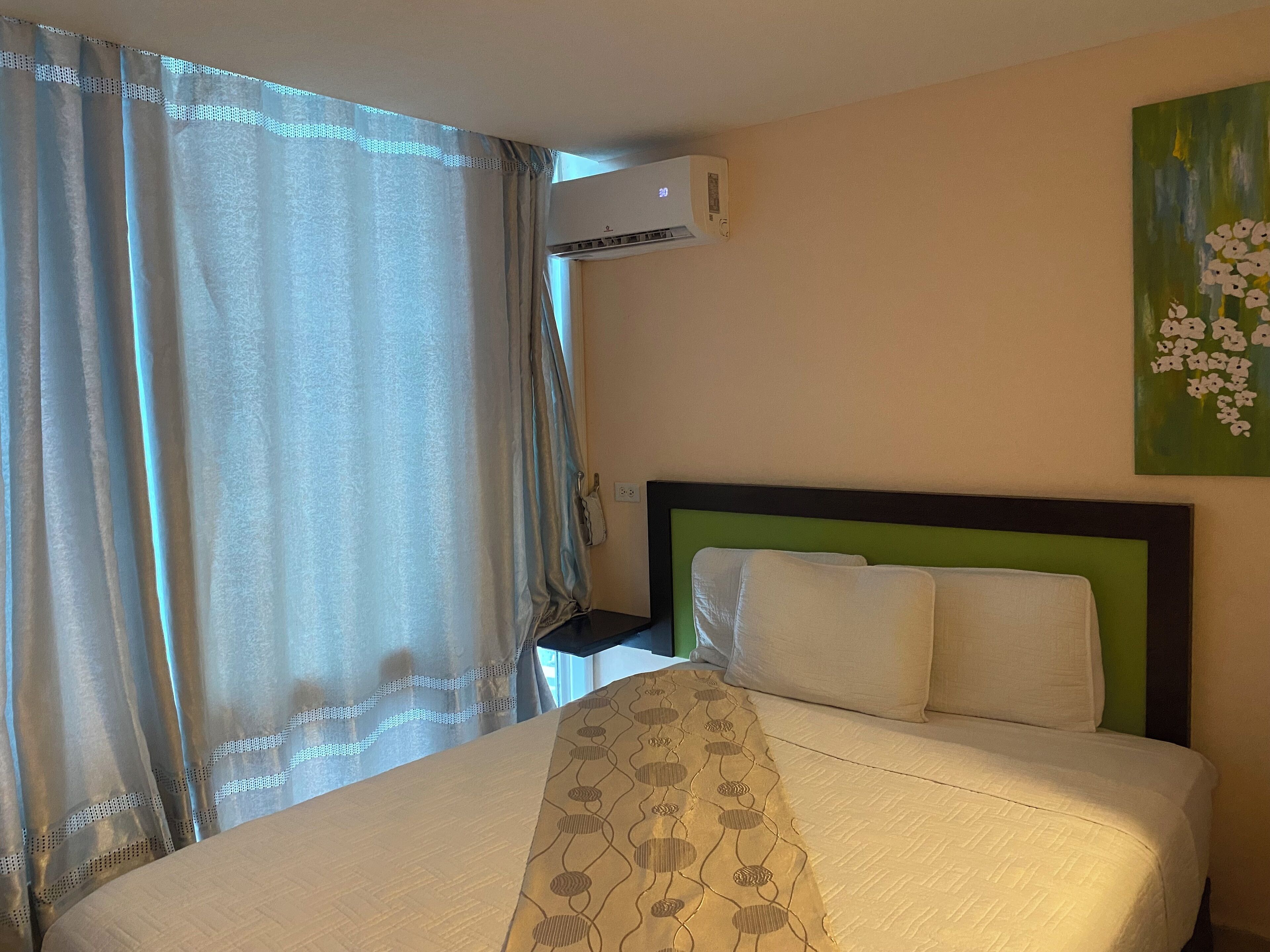 A hotel room with a bed, a wall-mounted air conditioner, and a painting of flowers.