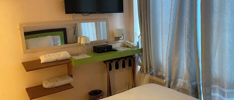 A hotel room with a bed, a desk with a telephone, a television, a mirror, and a window with curtains.