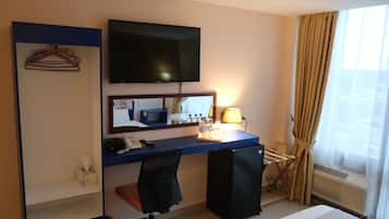 Habitación de hotel con escritorio, silla, cama, televisor y ventana con cortinas.