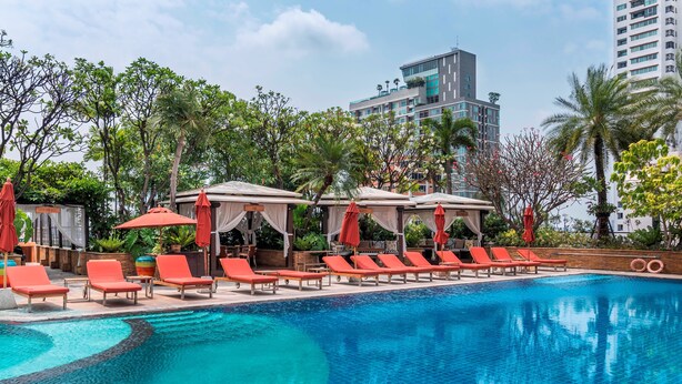Una piscina con sillas de descanso y sombrillas, rodeada de vegetación y edificios modernos.
