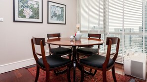 Un comedor con una mesa redonda, cuatro sillas, dos cuadros enmarcados con motivos florales, una ventana con persianas y un radiador.