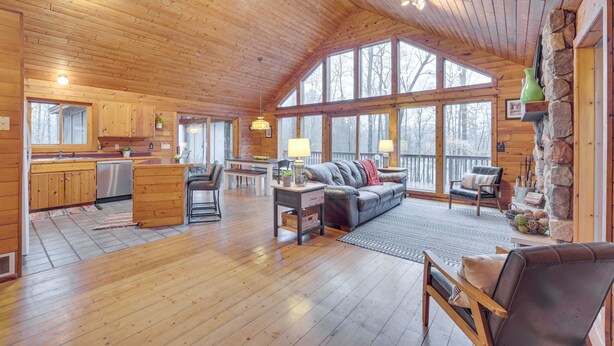 A spacious cabin with wooden floors, a large living area with a sofa and chairs, a kitchen with wooden cabinets, and large windows offering views of the surrounding forest. - Cozy Wisconsin Cabin w/ Deck, Kayaks & Lake Views! (Hayward)