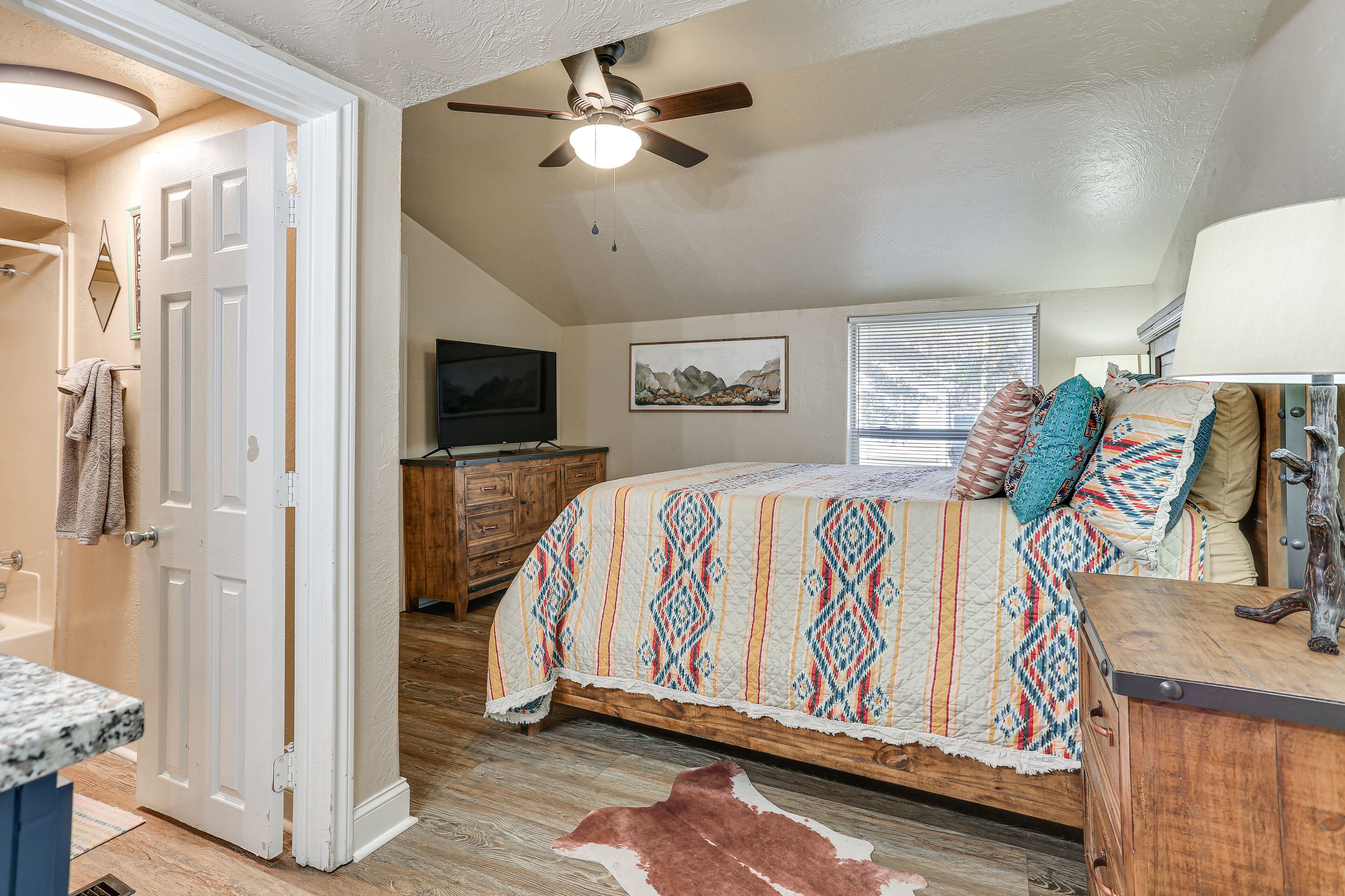 A bedroom with a bed, a ceiling fan, a wooden dresser, a television, and a window with blinds.