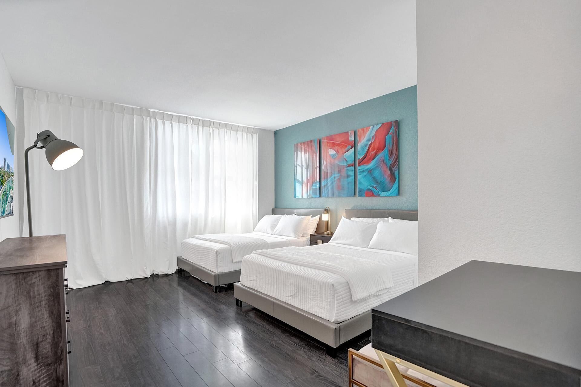 Hotelkamer met twee bedden, een groot abstract schilderij, een bureau met lamp en uitzicht op een stadsbeeld.