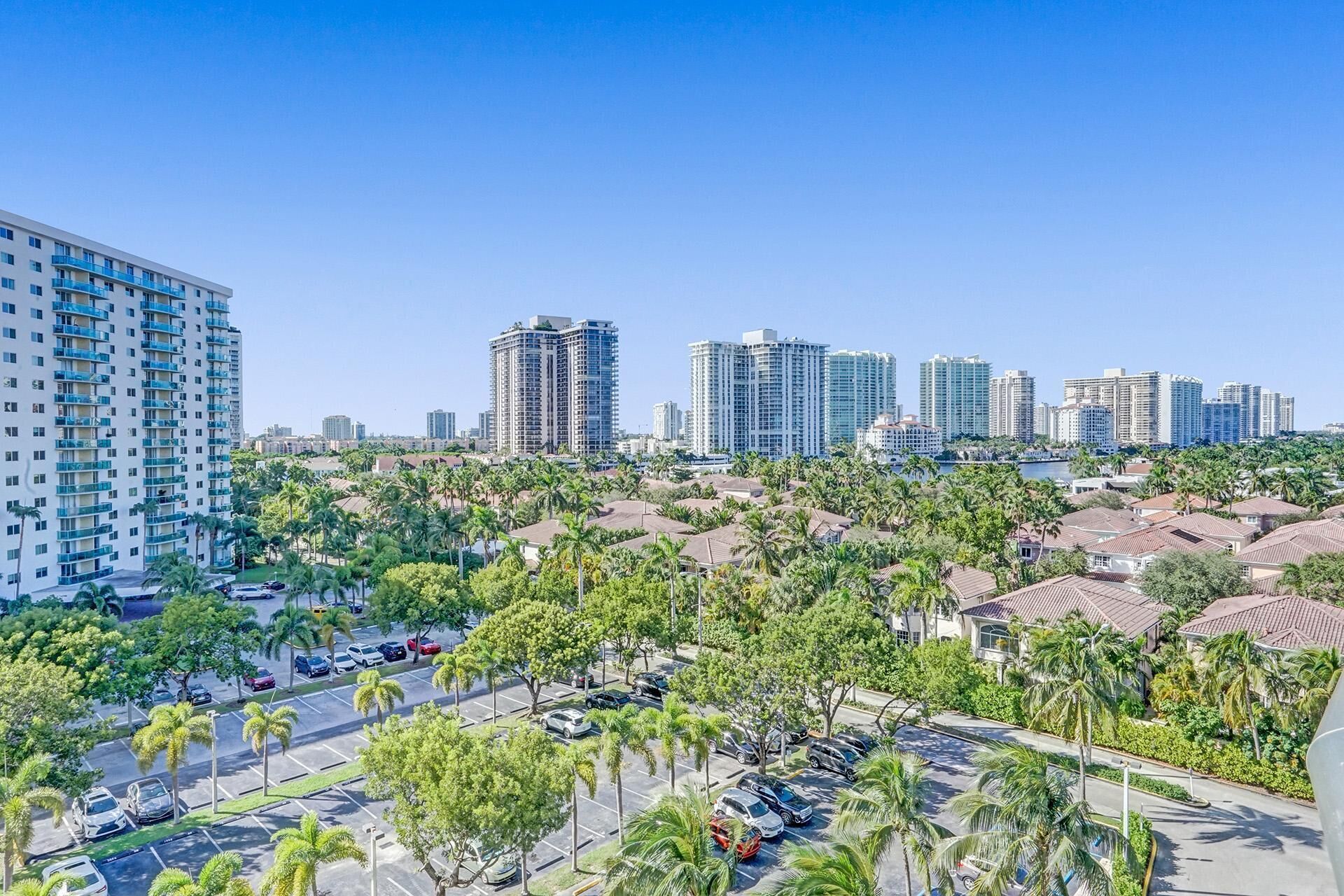 A high-rise building with multiple balconies, a parking lot with cars, and a clear blue sky.