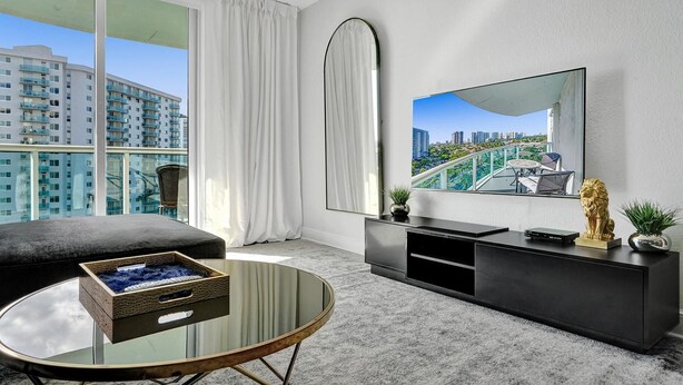 A modern living room with a large window, a round glass coffee table, a black sofa, a black entertainment unit, and a mounted TV.