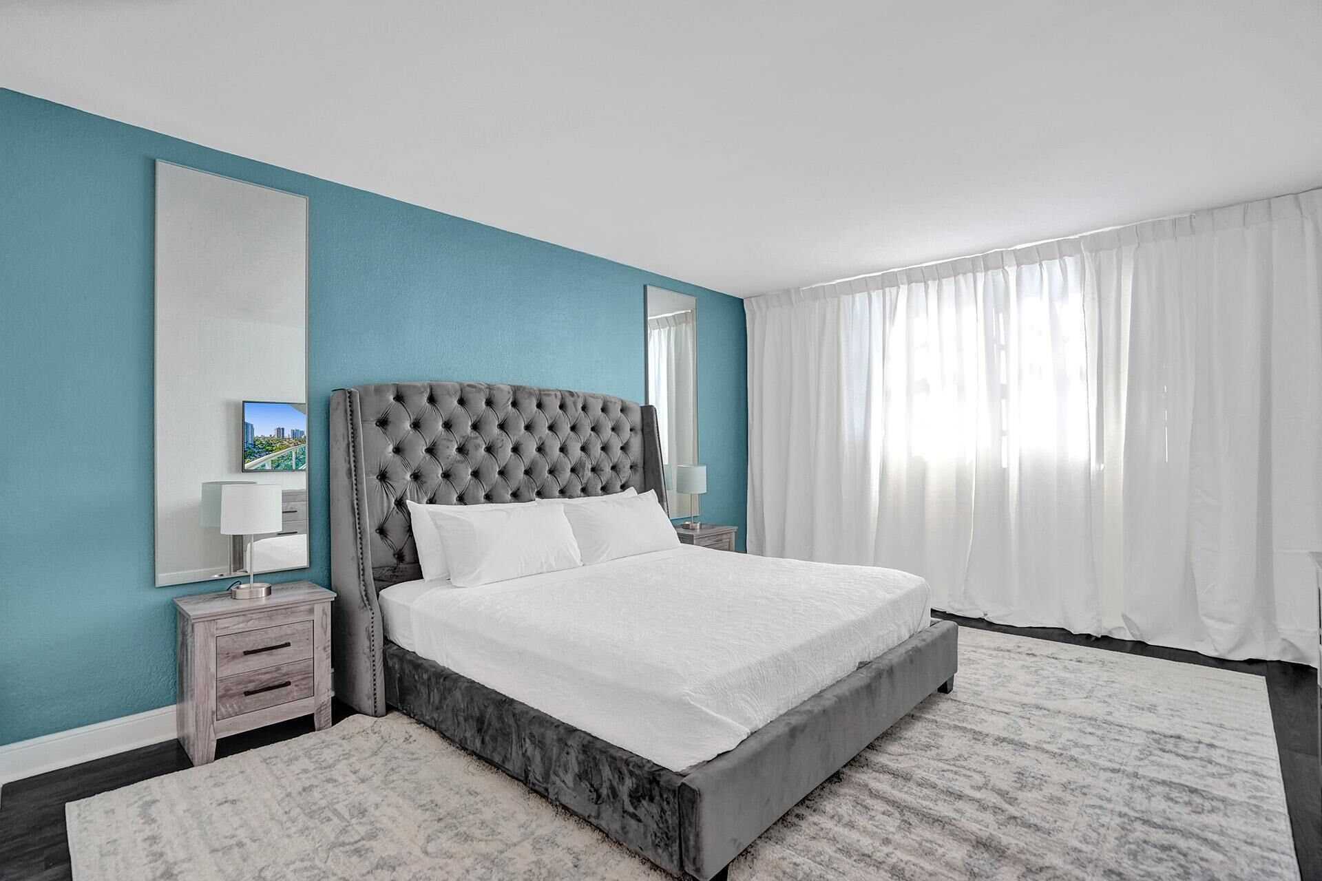 A modern hotel room with a large bed, a nightstand, a mirror, and a view of the outdoors through sheer curtains.
