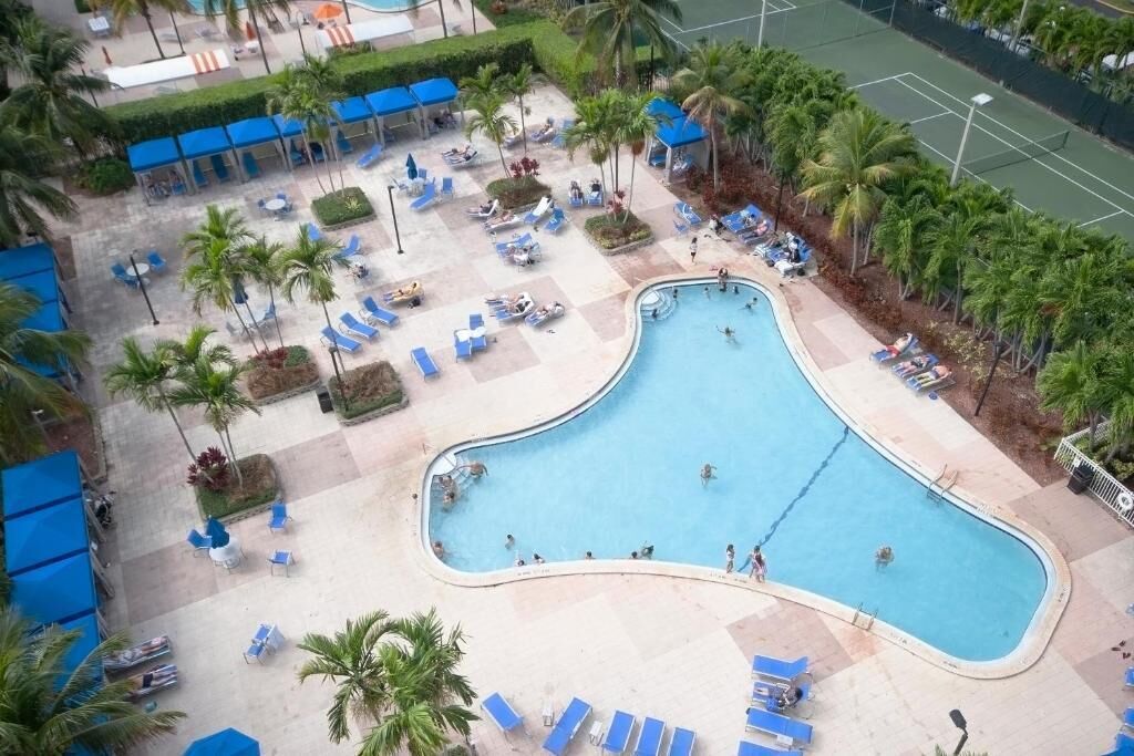 An aerial view of a resort with a large swimming pool surrounded by palm trees, lounge chairs, and a tennis court.