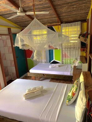 A room with two beds, a ceiling fan, a mosquito net, and a small table with a bottle. - Le Petit Paradis (Roseau)