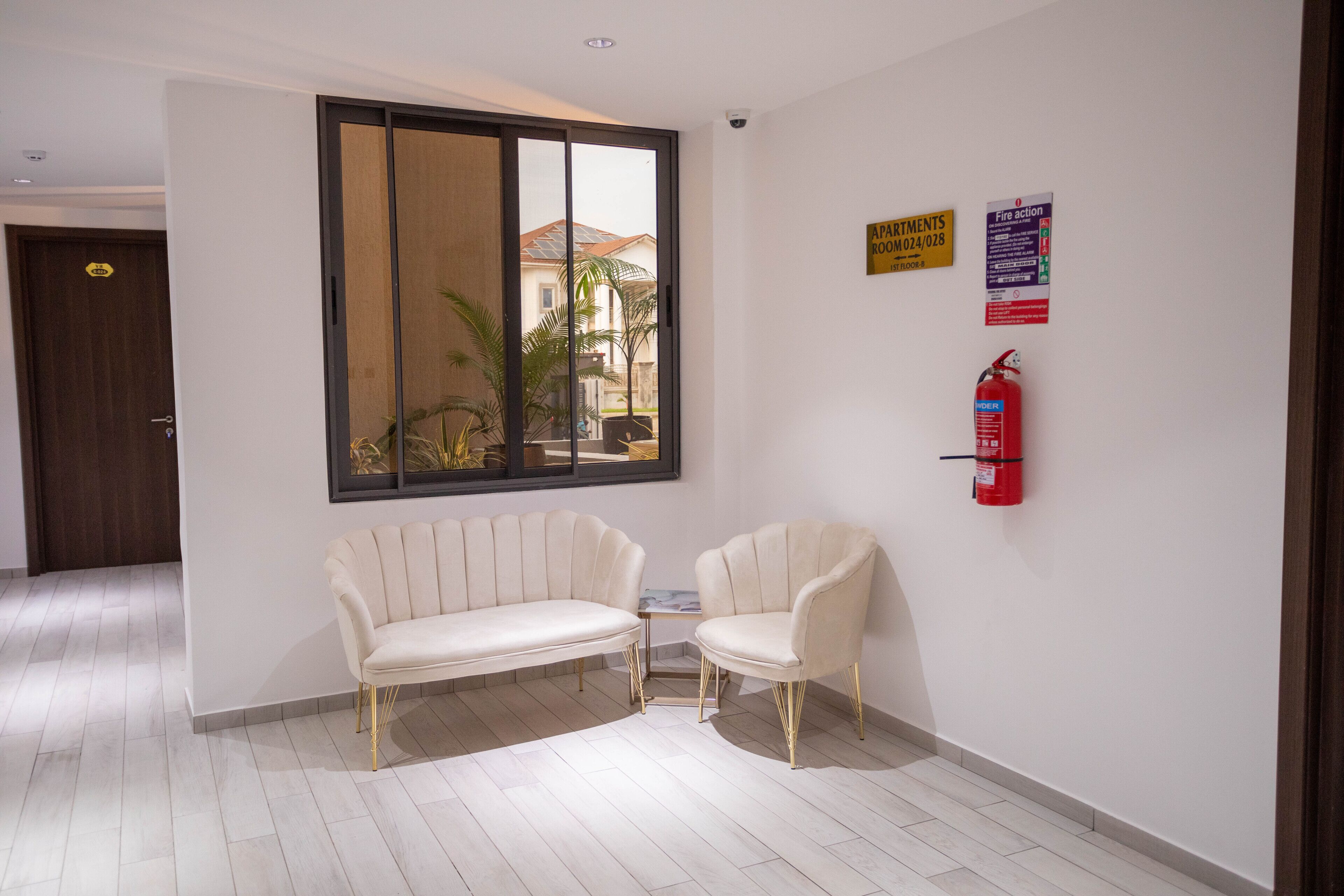 A hotel lobby with a sofa and chairs, a fire extinguisher, and a window with a view of a building and palm trees.
