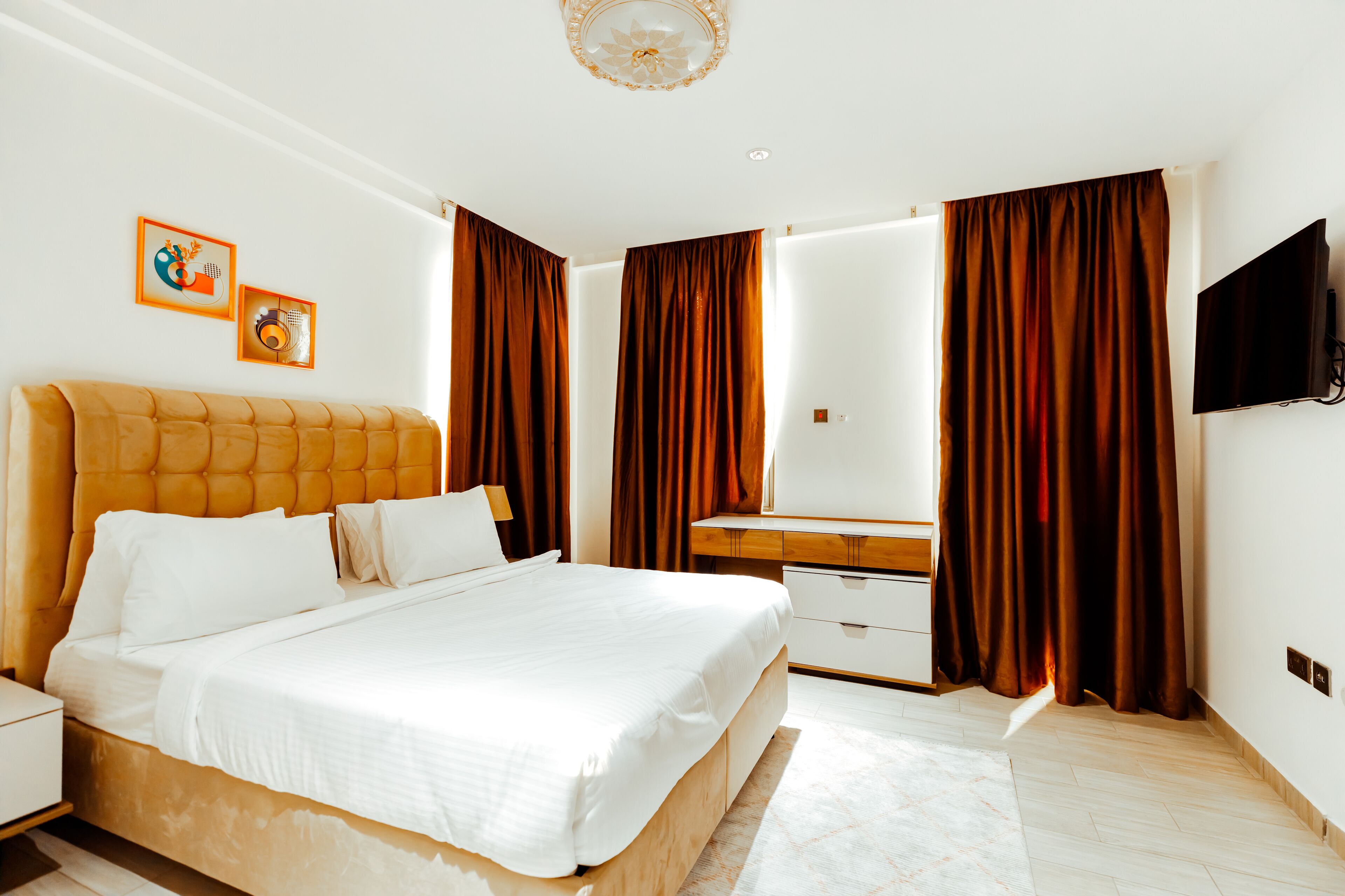 A modern hotel room with a large bed, a wooden nightstand, a dresser, a flat-screen TV, and two framed pictures on the wall.