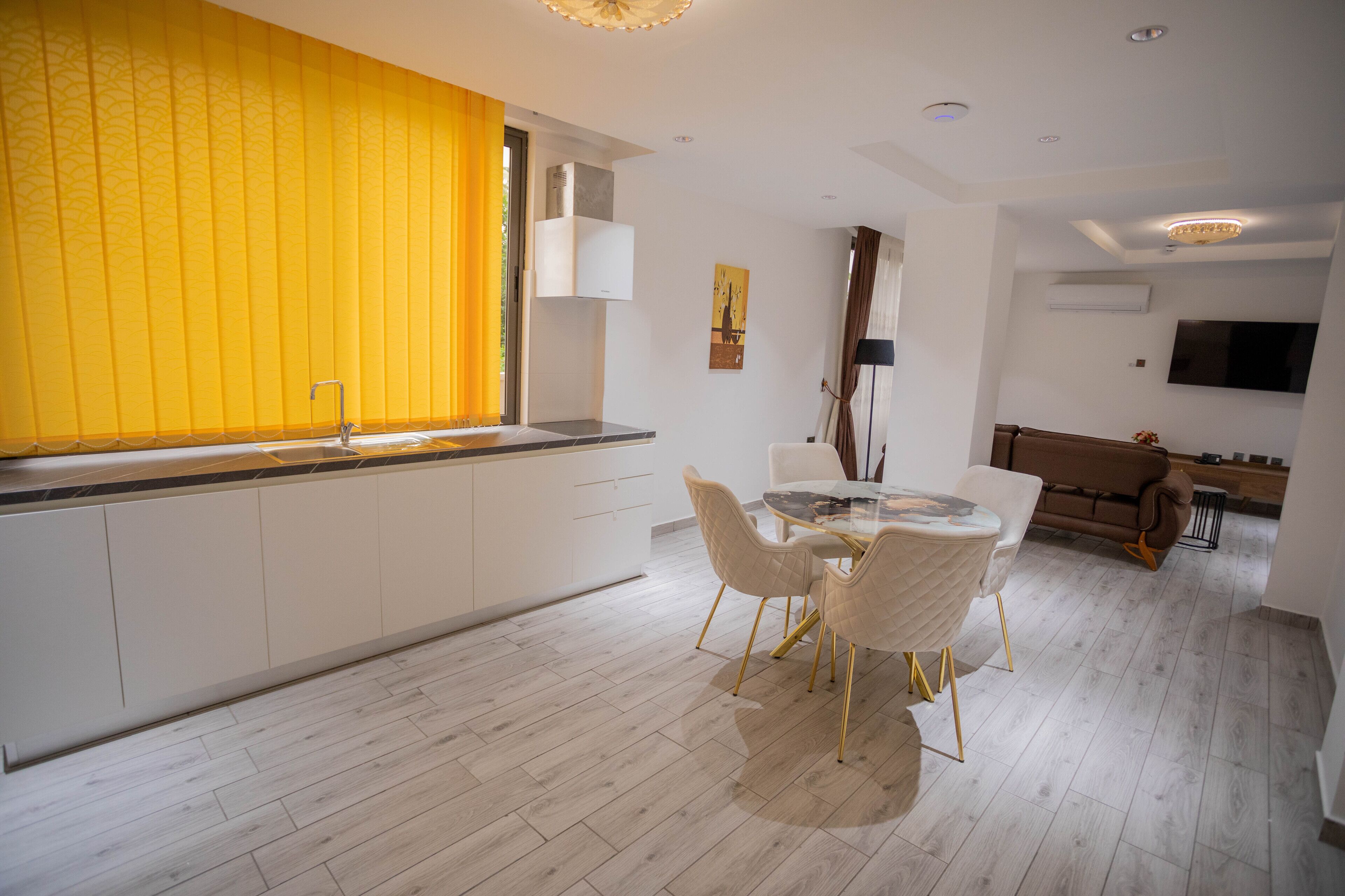 A modern kitchen with white cabinets, a yellow window blind, and a dining area with a glass table and white chairs.
