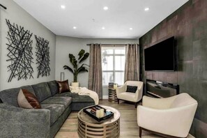 Una sala de estar moderna con un sofá gris, un sillón blanco, una mesa de centro redonda y un televisor de pantalla plana montado en la pared.