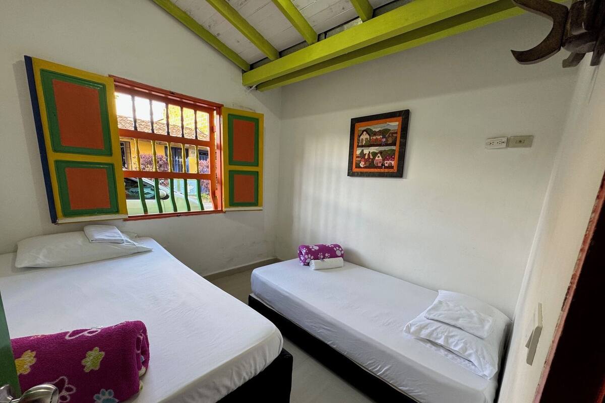 A room with two single beds, a window with colorful shutters, a painting on the wall, and a door with a metal handle.