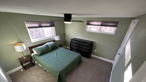 Une chambre à coucher comprenant un lit, une commode, une table de chevet, un ventilateur de plafond et deux fenêtres équipées de stores.