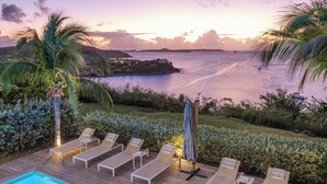 Un espace aménagé au bord de la piscine, avec des chaises longues et un parasol, offrant une vue sur une étendue d’eau au coucher du soleil.