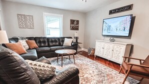 A living room with a black leather sofa, a wooden coffee table, a patterned rug, a white cabinet, and a flat-screen TV mounted on the wall.