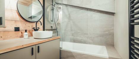 Un baño moderno con un tocador de madera, un espejo redondo, un lavabo blanco y una ducha con mampara de vidrio.