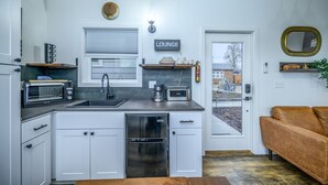 Cocina compacta con armarios blancos, horno empotrado, un lavamanos y cafetera. Un cartel sobre la puerta indica “LOUNGE”, y hay un espejo en la pared.
