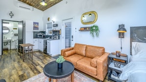 Un salón espacioso con un sofá de cuero marrón, una mesita de centro redonda y una mesita auxiliar con lámpara. Se ve una cocina al fondo, con una zona de comedor y una puerta que da a un baño.