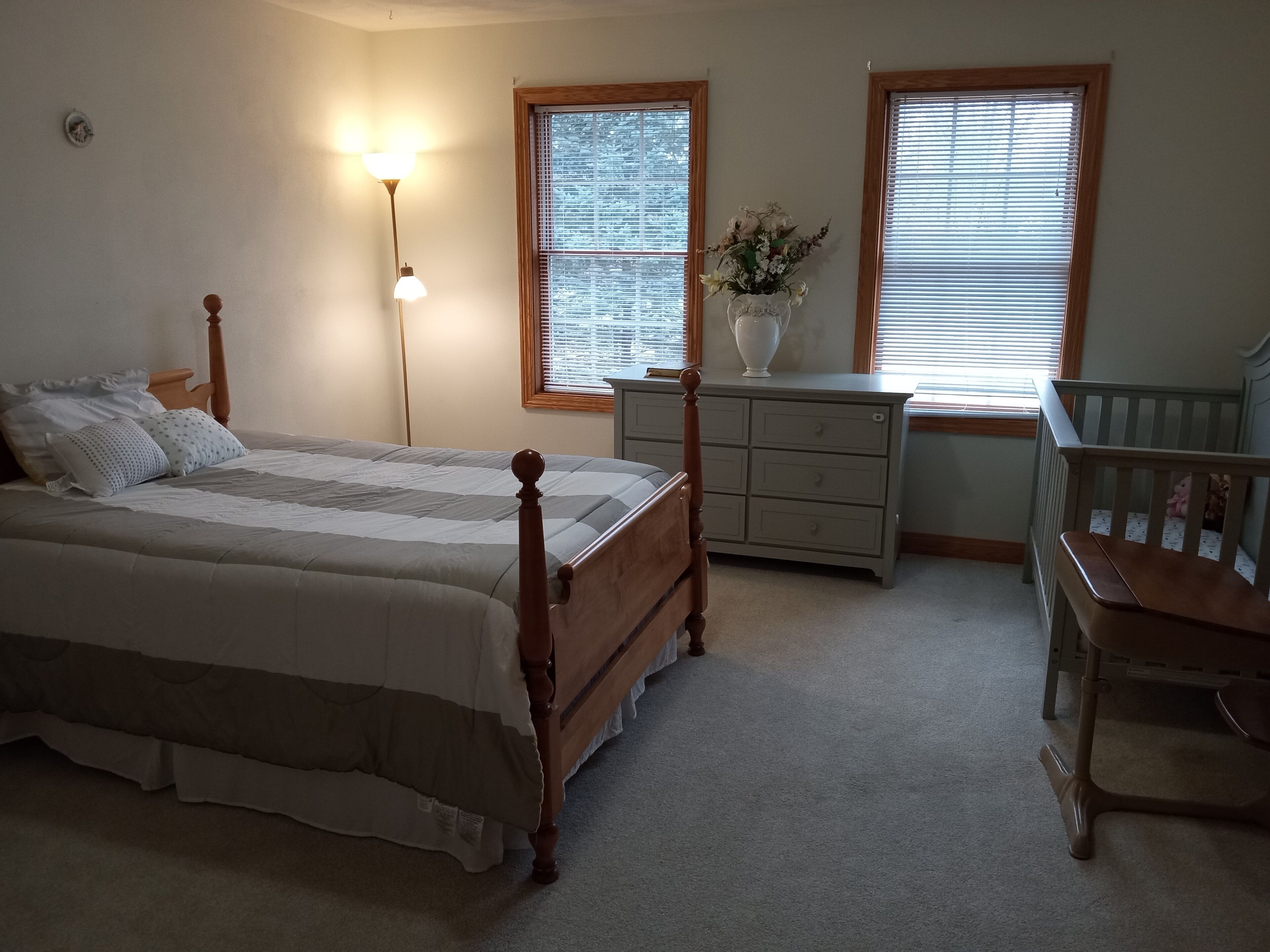 A bedroom with a bed, dresser, nightstand, lamp, windows, and a crib.