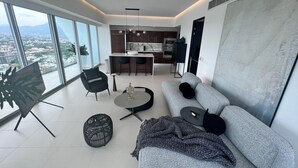Una sala de estar moderna con un sofá gris, una mesa de centro y una silla negra. Se observa una cocina al fondo con una zona de comedor.
