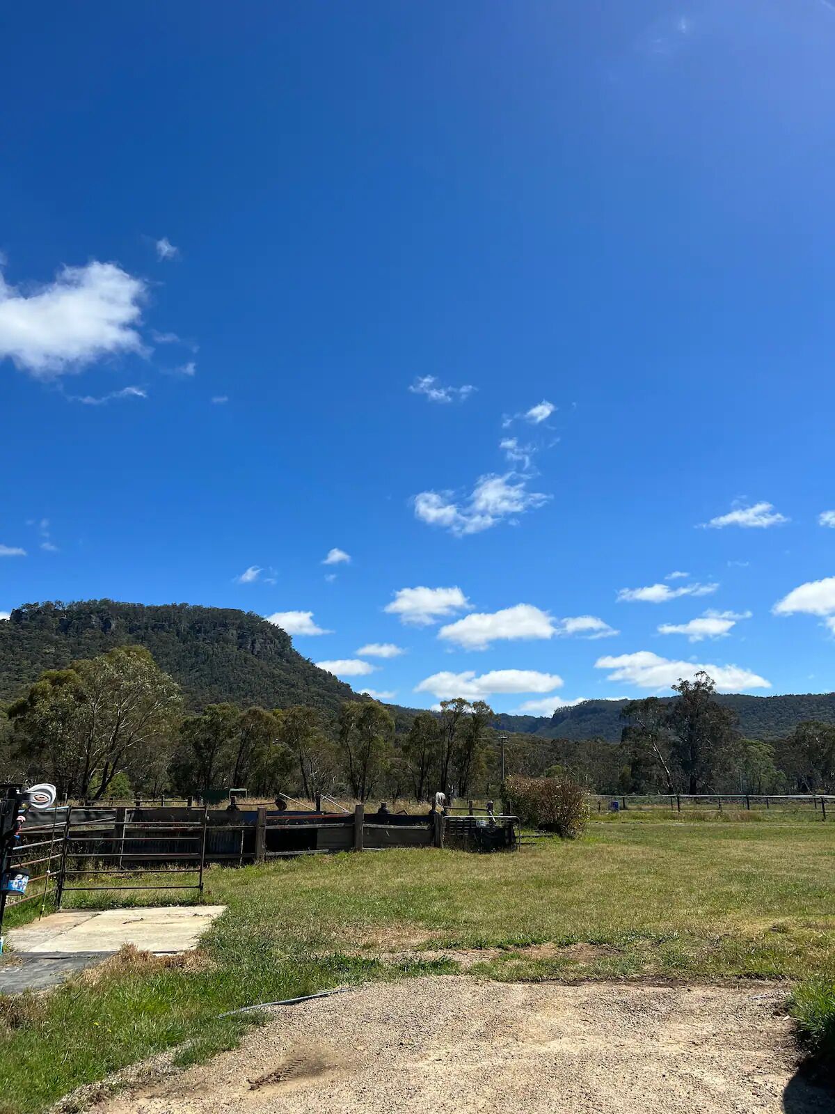A rural landscape with a clear blue sky, a dirt path, and a fenced area with trees and a hill in the background.