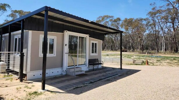 A small, single-story building with a covered porch, large sliding glass doors, and a bench outside. - Oliver’s Hut - Hartley Huts (Hartley Vale)