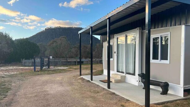 A modern building with a covered porch and large windows, set against a backdrop of a fenced area and hills. - Yabba’s Hut - Hartley Huts (Hartley Vale)